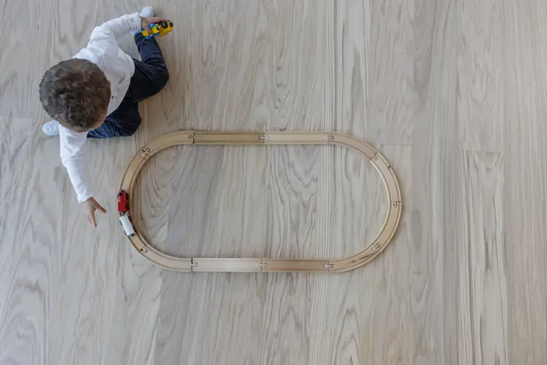 Child playing with a wooden train set on a light wood floor, wearing a white shirt and blue pants.