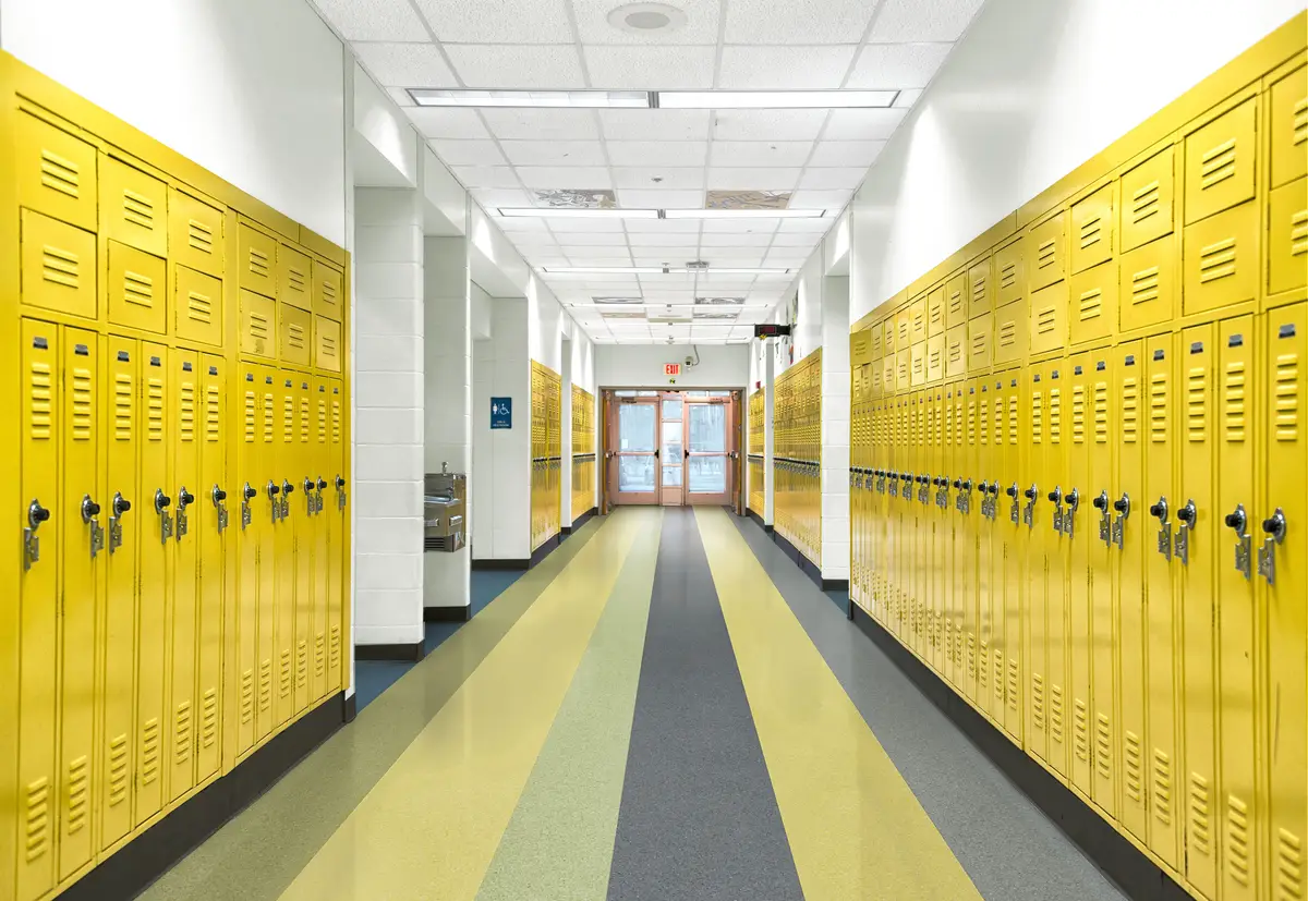 A school hallway with rows of yellow lockers on both sides, a striped floor, and bright overhead lighting leading to double doors.