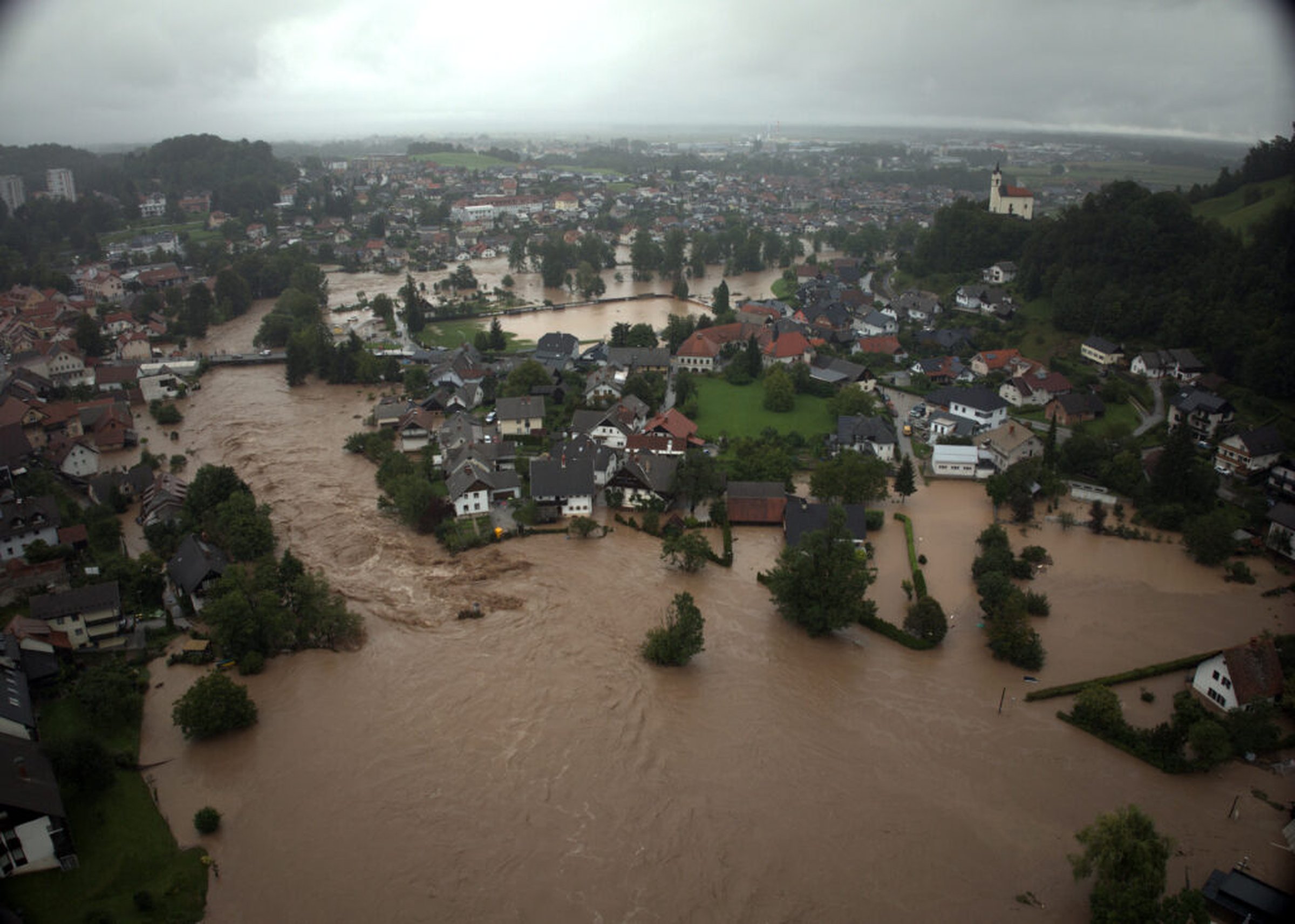 Aerial view of a town flooded by muddy waters, with submerged roads and partially inundated houses under a cloudy sky.