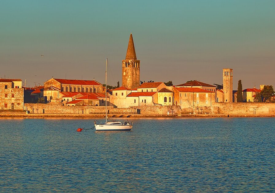 Coastal town at sunset with historic buildings, a prominent tower, and a sailboat on calm water.