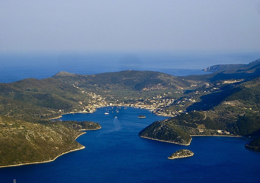 Aerial view of a coastal town surrounded by lush hills and a deep blue sea, with a narrow inlet leading to a harbor.