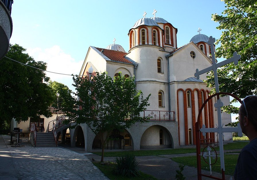 A white church with red accents and multiple domes, surrounded by trees and a paved pathway in a sunny setting.