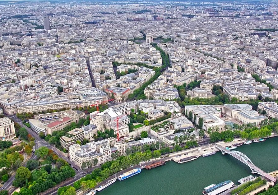 Aerial view of Paris showcasing the Seine River, numerous buildings, and green spaces under a cloudy sky.
