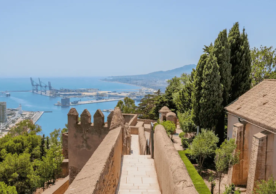 View from a stone pathway overlooking a coastal city with a harbor, surrounded by trees and historic architecture under a clear blue sky.
