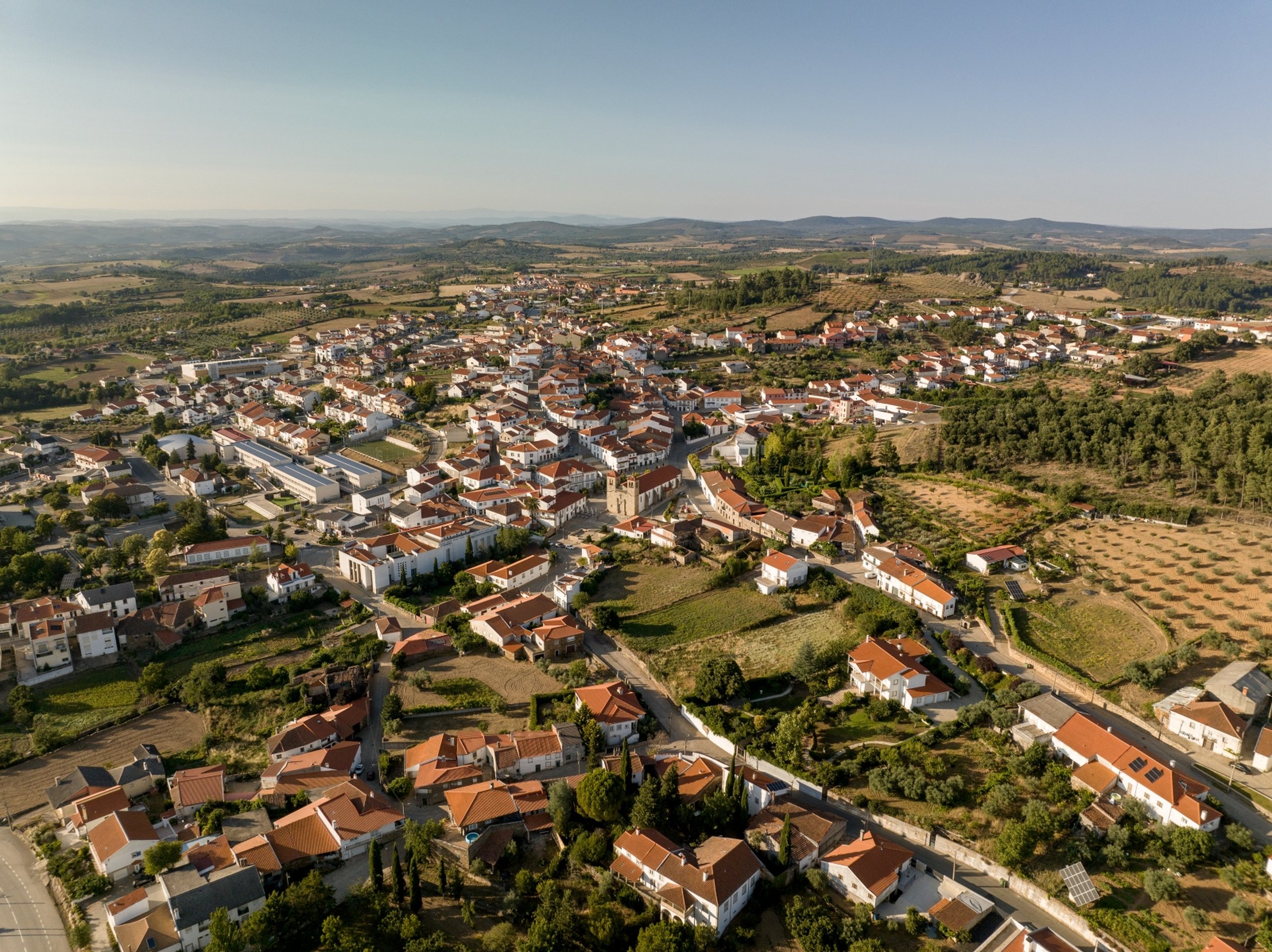 Aerial view of a picturesque town with red-roofed buildings, surrounded by fields and hills under a clear blue sky.