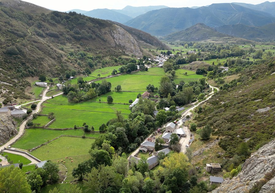 Aerial view of a lush valley with green fields, scattered houses, winding roads, and surrounding hills under a cloudy sky.