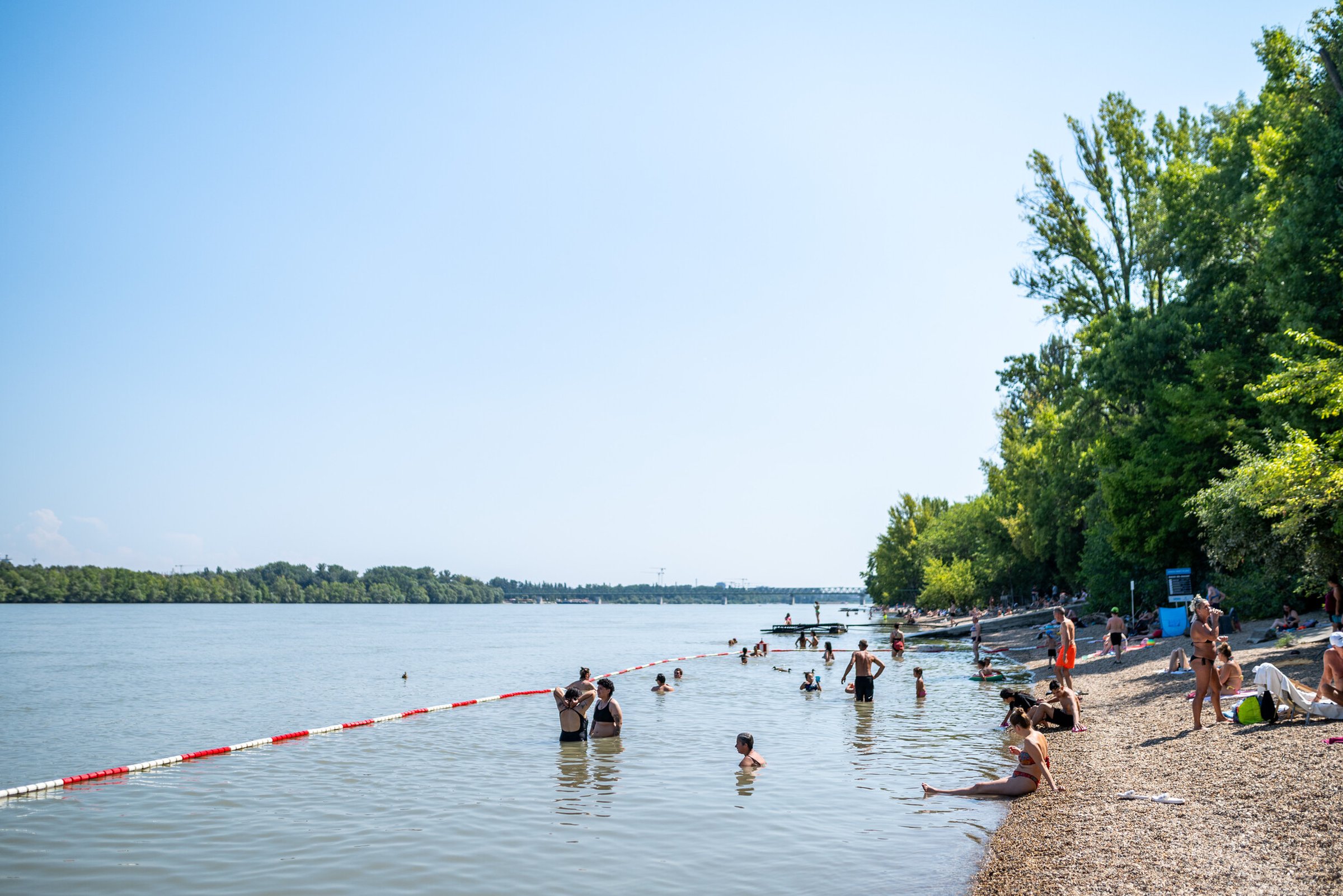 People swimming and relaxing on a pebble beach by a calm river, with trees lining the shore under a clear blue sky.