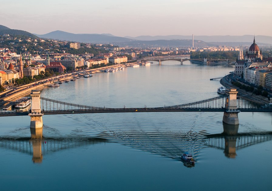 Aerial view of a bridge over a wide river, with a boat passing underneath. Cityscape and distant hills visible under a clear sky.