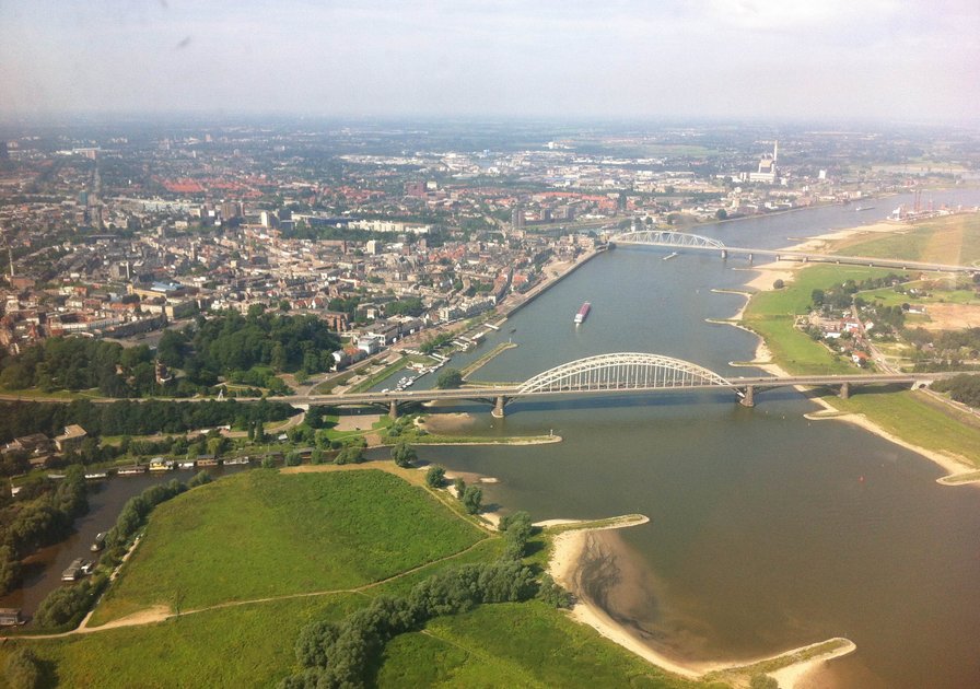 Aerial view of a city with two bridges spanning a river, surrounded by green fields and urban buildings under a clear sky.
