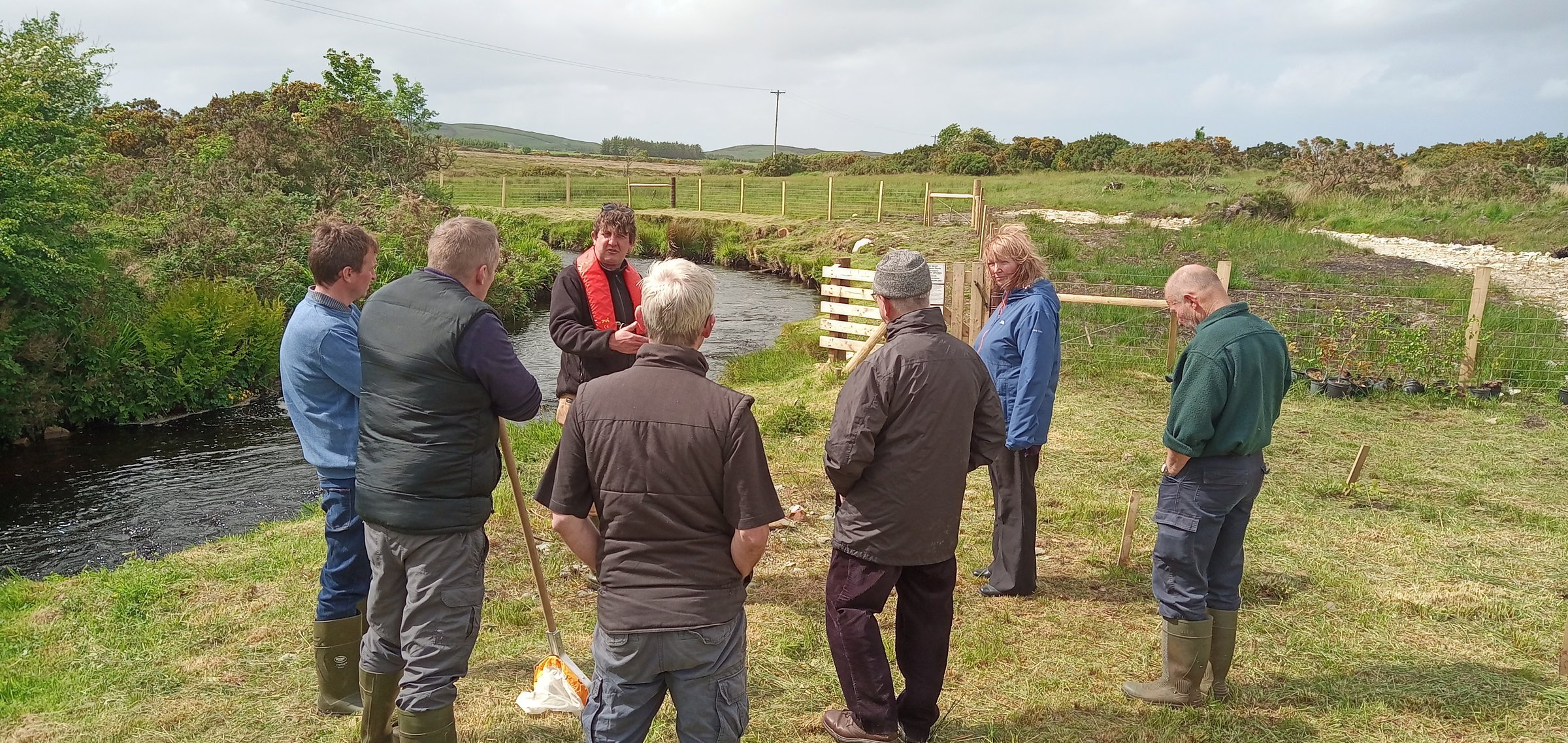 A group of people stands in a grassy field near a small stream, engaged in conversation. Some are wearing boots and jackets.