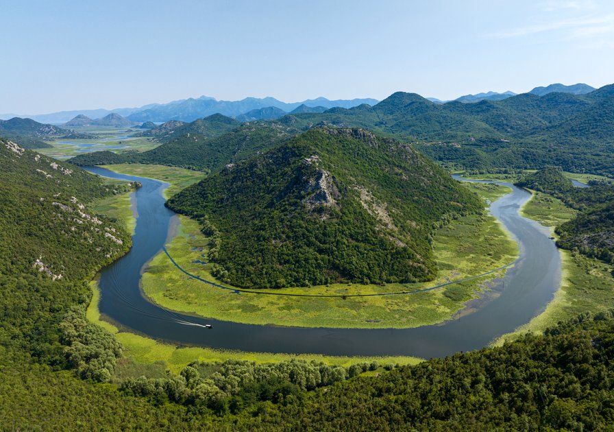 Aerial view of a winding river encircling a lush, green hill with mountains in the background under a clear blue sky.