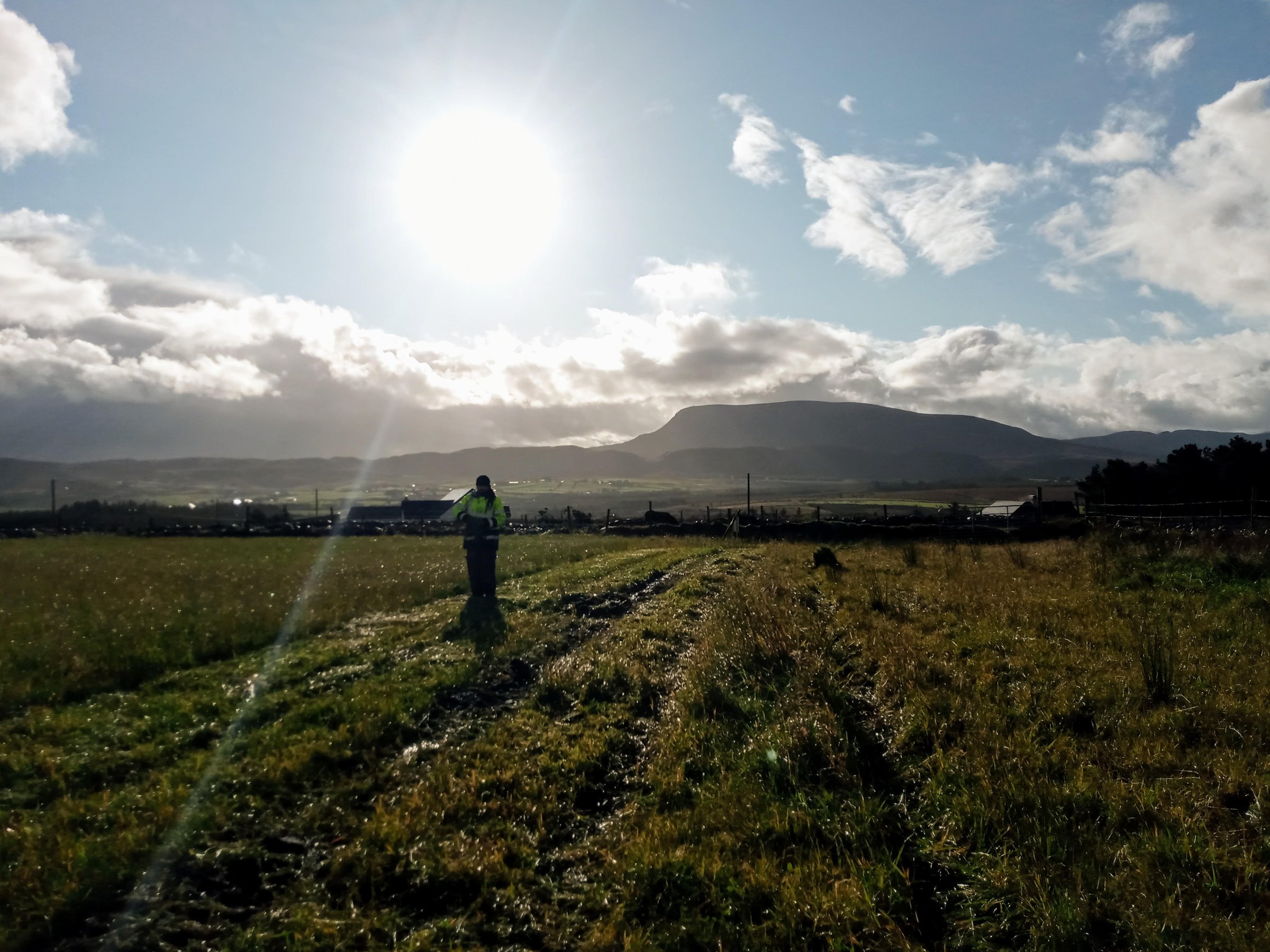 A person in a field under a bright sun, with distant mountains and scattered clouds in the background.