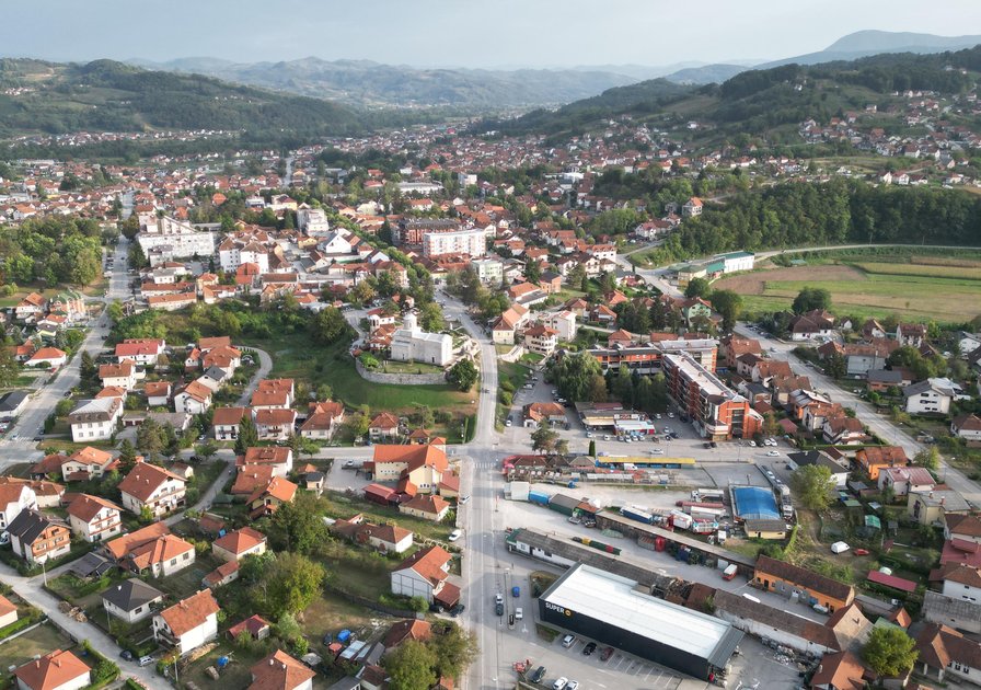 Aerial view of a town with red-roofed buildings, roads, and surrounding green hills under a cloudy sky.