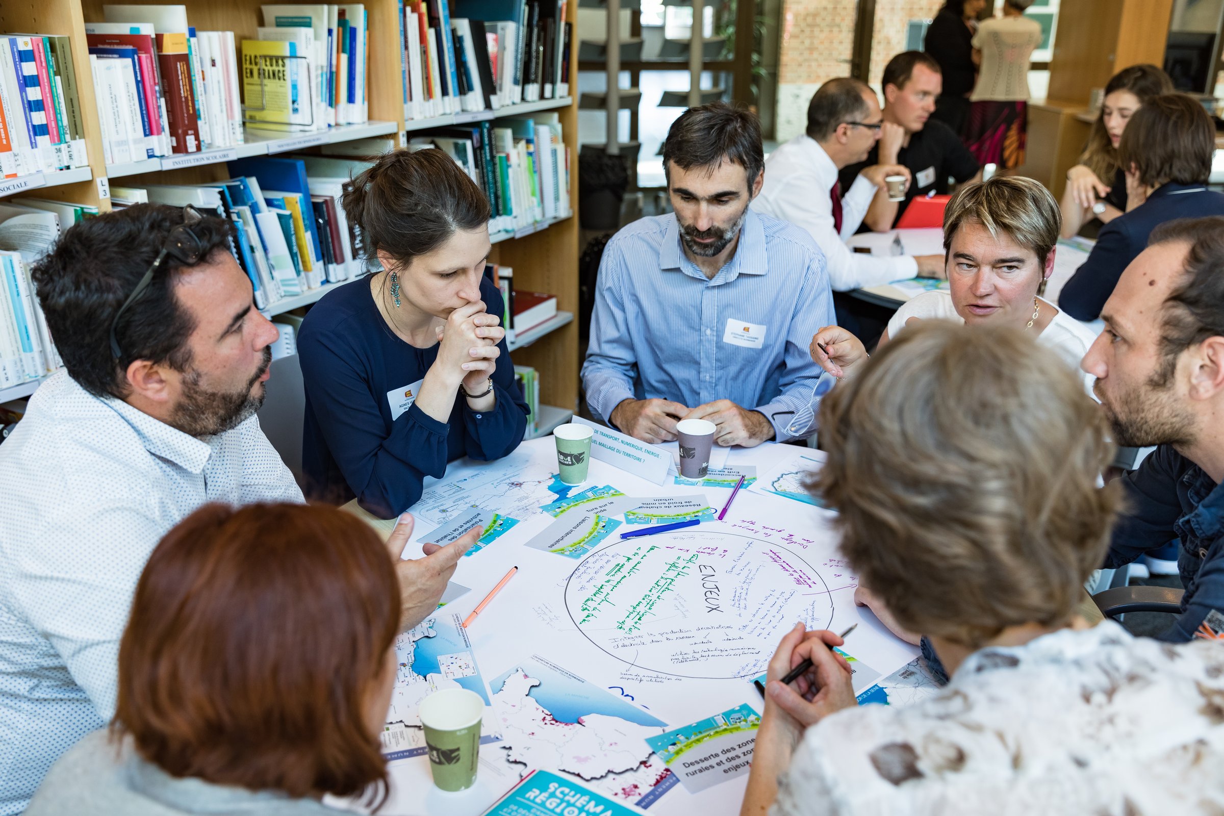 A group of people engaged in discussion around a table covered with papers and markers in a library setting.