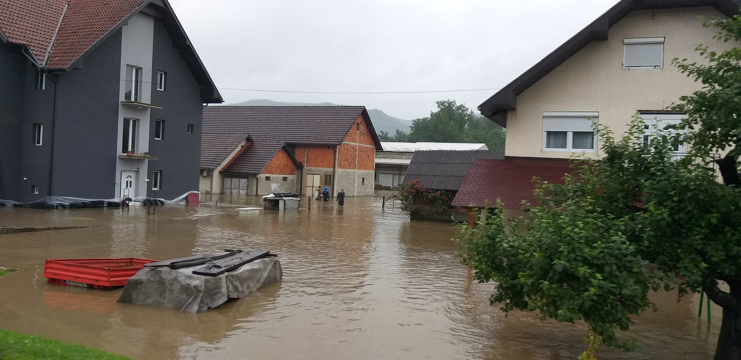Flooded residential area with partially submerged houses, trees, and a red boat. Overcast sky and murky water covering the ground.