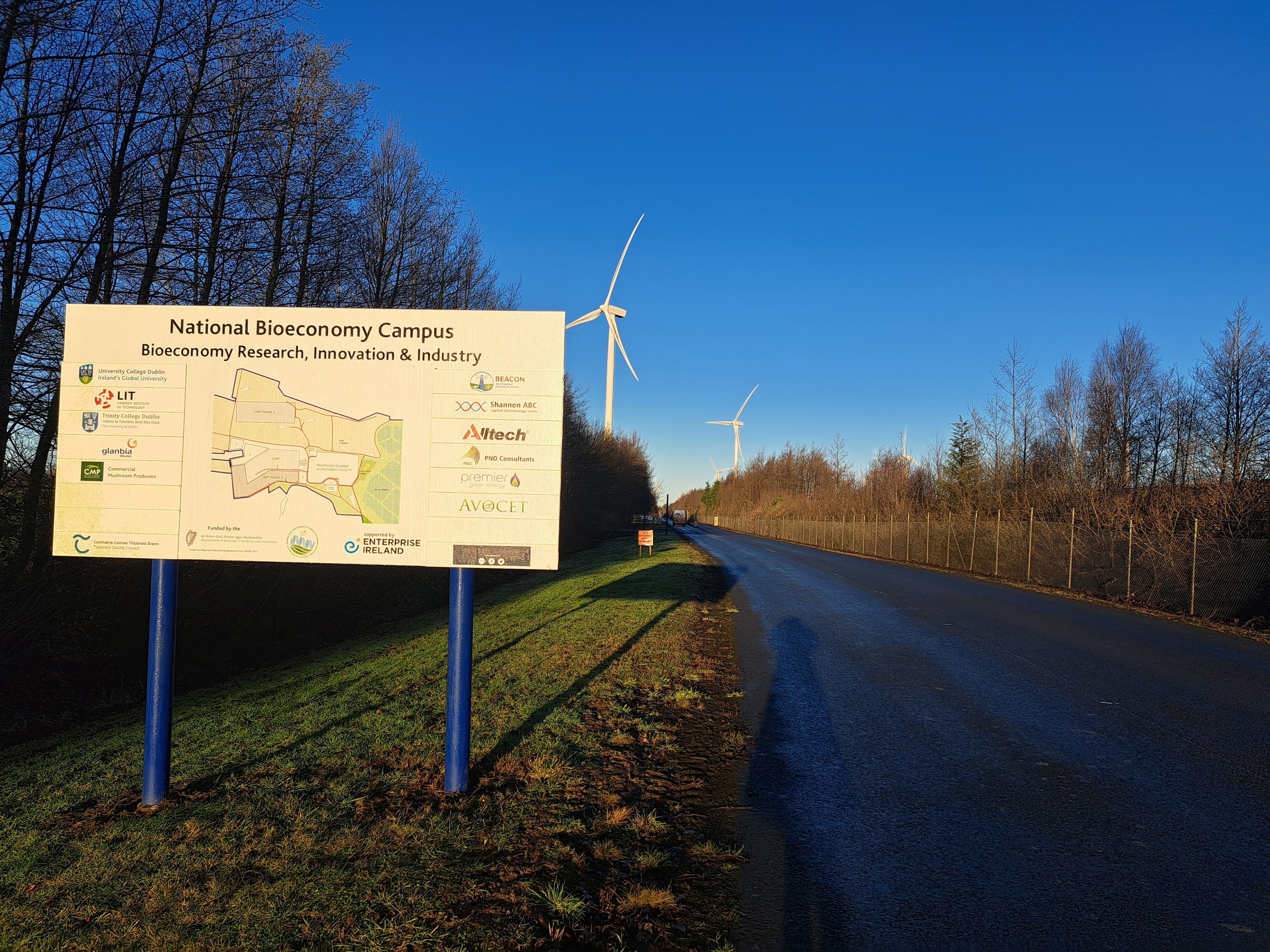 Sign for the National Bioeconomy Campus beside a road with wind turbines and trees under a clear blue sky.