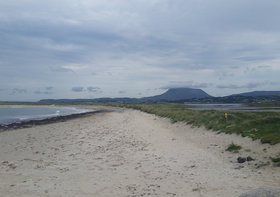 Sandy beach with grassy dunes under a cloudy sky, distant mountains visible on the horizon.