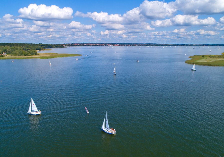 Aerial view of a calm lake with sailboats scattered across the water, surrounded by greenery and under a sky filled with fluffy clouds.