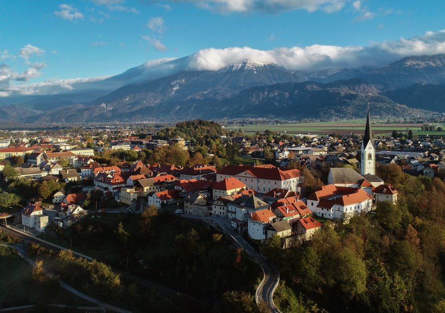Aerial view of a picturesque town with red-roofed buildings, a church, and mountains in the background under a cloudy blue sky.