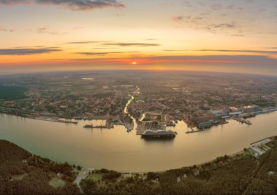 Aerial view of a coastal city at sunset, featuring a river winding through urban areas and a large ship docked near the shore.