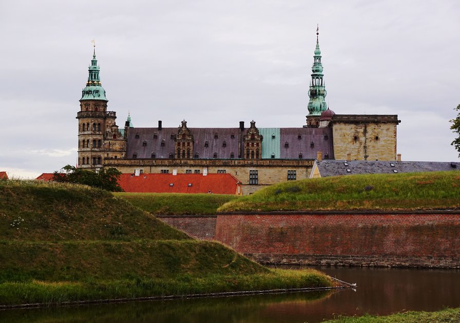 Kronborg Castle in Denmark, with its spires and towers, stands behind grassy embankments and a moat under a cloudy sky.