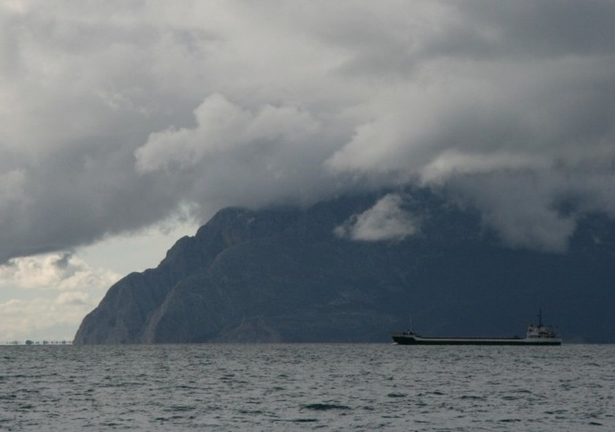 A cargo ship sails on a vast body of water under a cloudy sky, with a mountainous landscape in the background.