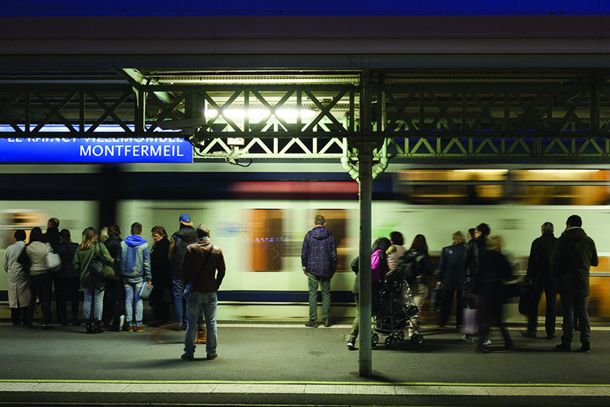 People wait on a dimly lit train platform labeled "Montfermeil" as a train speeds by, creating a motion blur effect.