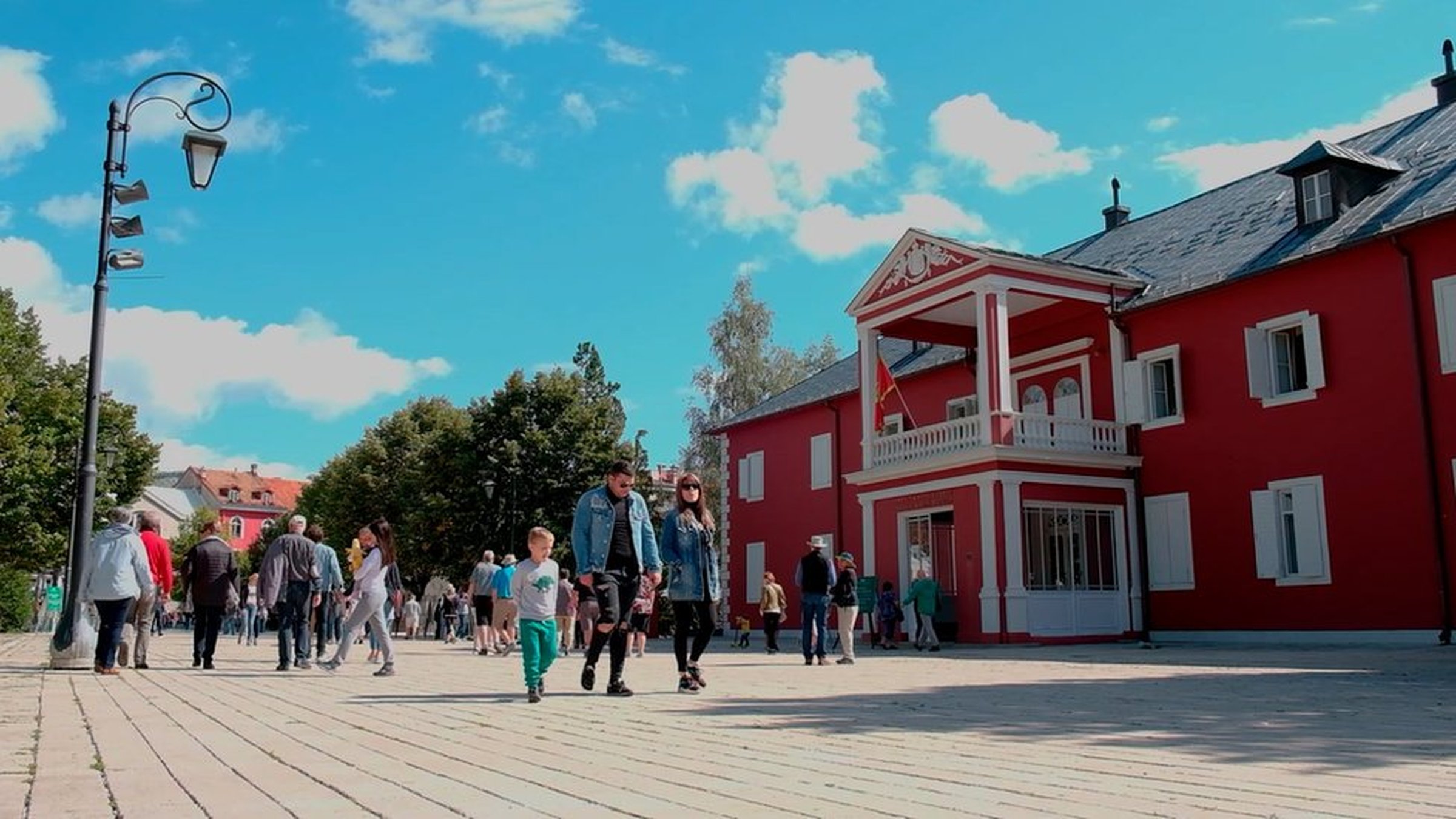 People walking on a sunny day near a red building with white trim and a balcony, surrounded by trees and a clear blue sky.