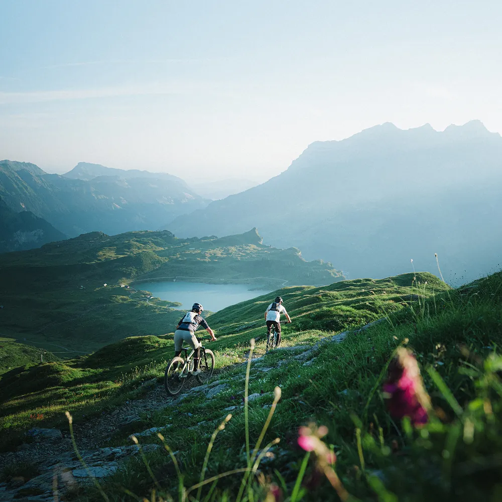 Zwei Mountainbiker fahren einen Trail hinunter mit Blick auf einen Bergsee