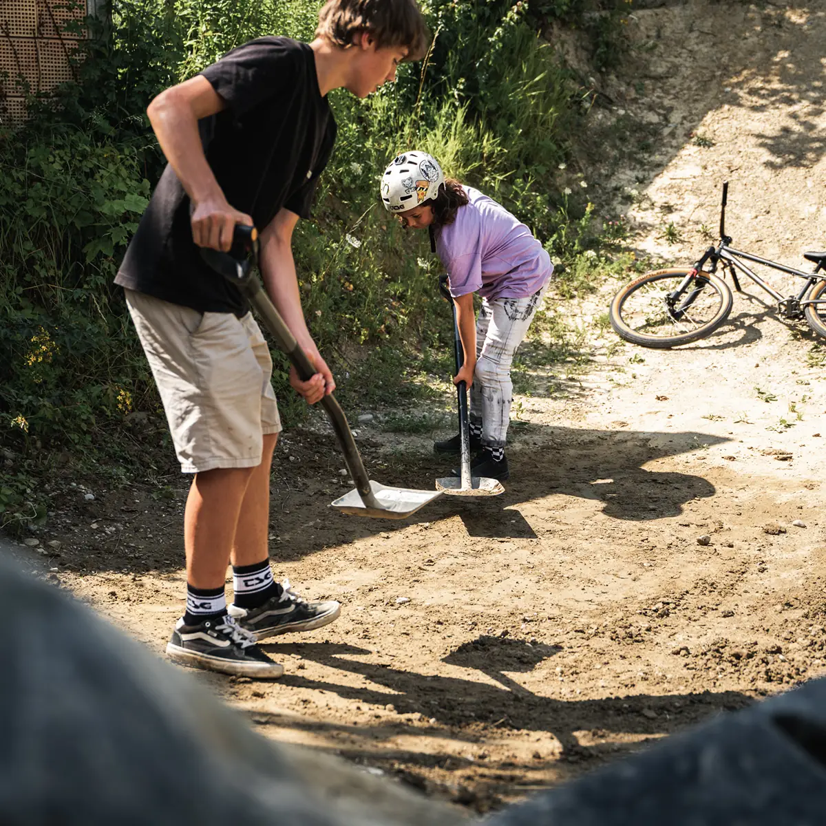 Zwei Jugendliche mit Helmen schaufeln einen Dirt-Trail, im Hintergrund ein BIXS Dirtjump-Bike