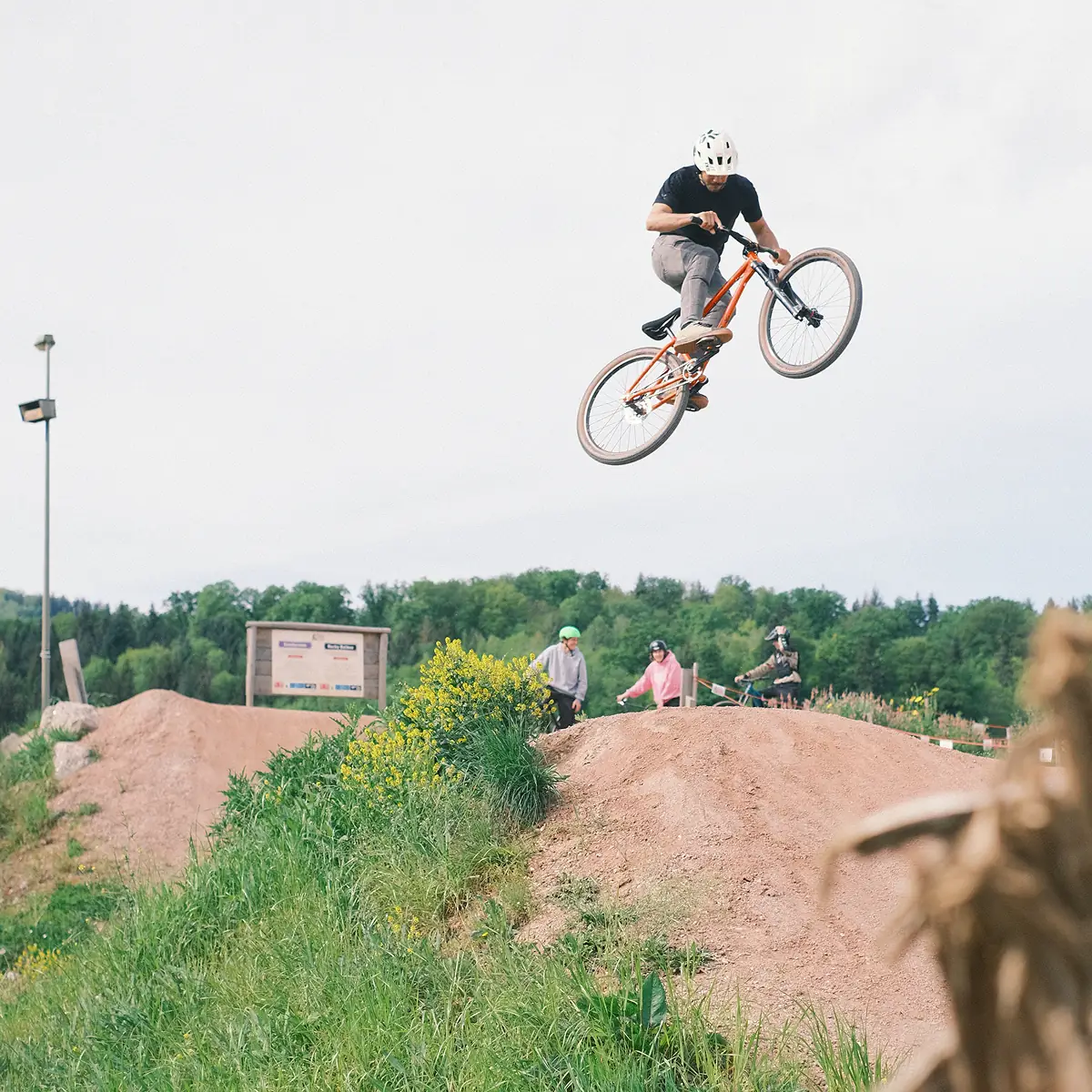 Fahrer springt mit orangem BIXS Dirtjump-Bike über einen Erdhügel, Zuschauer im Hintergrund