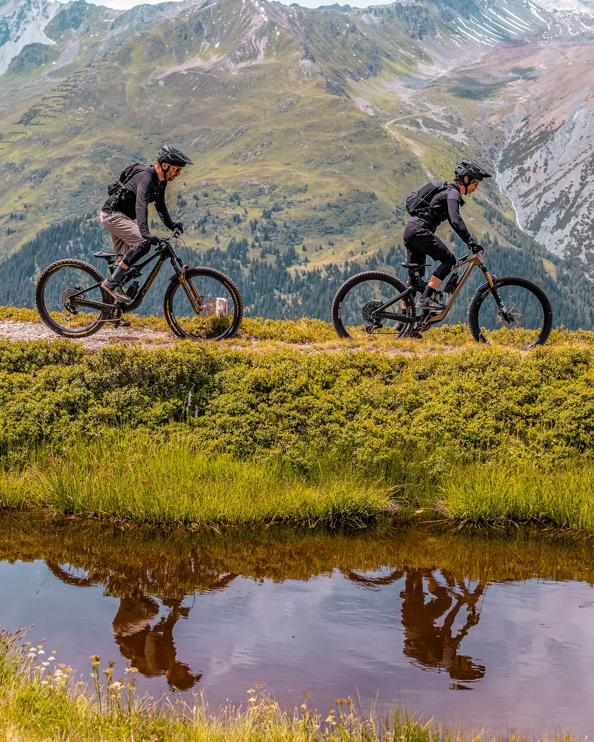 Zwei Fahrer auf Höhenweg mit Spiegelung in kleinem Bergsee