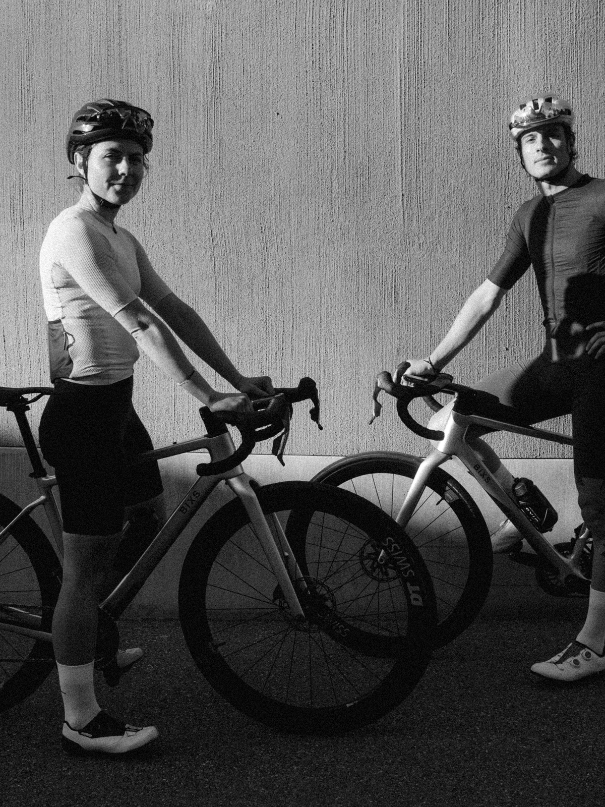 Zwei Rennradfahrer in Schwarzweiss posieren mit ihren Gravelbikes vor einer strukturierten Betonwand