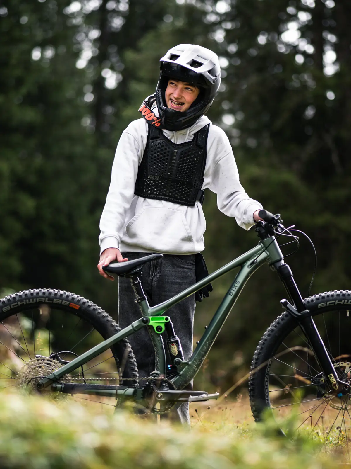 Martin mit Fullface-Helm und Protektor steht lächelnd neben seinem grünen BIXS Kids-Mountainbike im Wald
