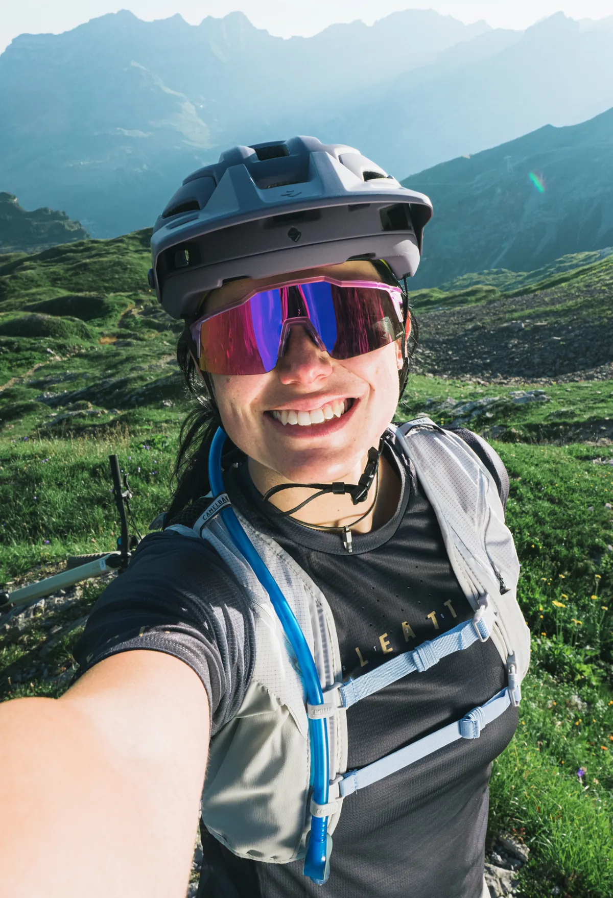 Mountainbikerin mit Helm und verspiegelter Brille macht ein Selfie auf einer Alpwiese vor Bergpanorama
