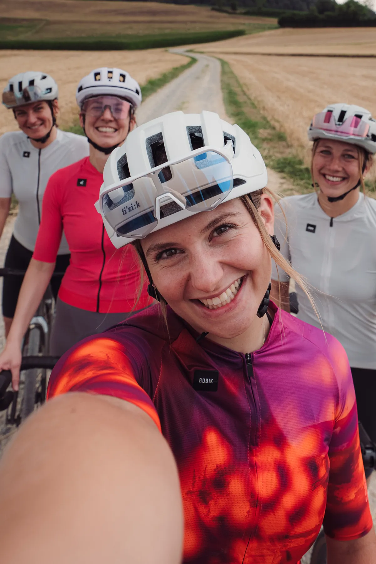 Vier Rennradfahrerinnen machen ein Selfie auf einem Kiesweg zwischen abgeernteten Feldern