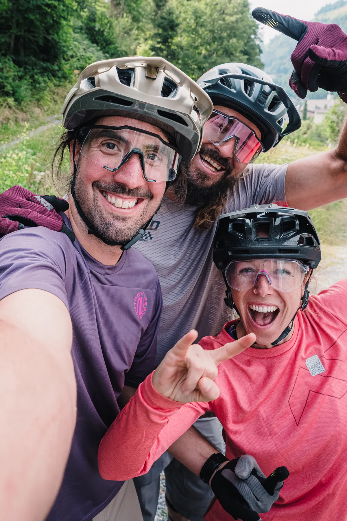 Drei Mountainbiker in Helm und Brille machen lachend ein Gruppenselfie am Waldrand