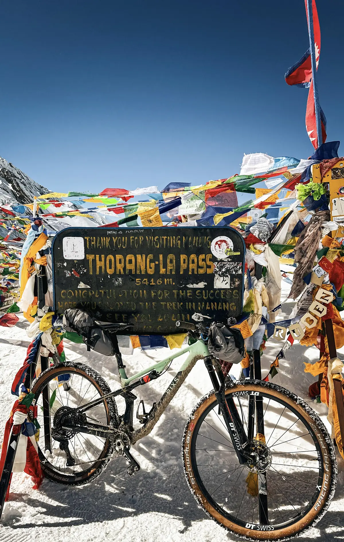 Bepacktes Bike lehnt an Gipfelschild "Thorong La Pass 5416m" mit Gebetsfahnen