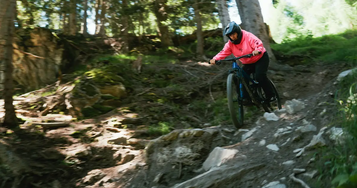 Fahrerin in pinkem Hoodie springt über Felsstufe im Waldtrail