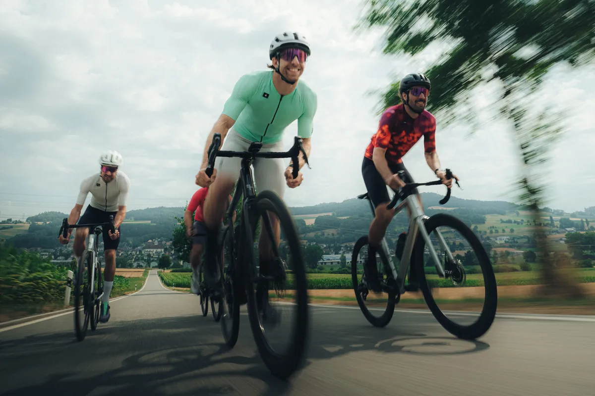 Vier Rennvelofahrer fahren lachend auf einer Landstrasse zwischen Feldern, im Hintergrund ein Dorf und bewaldete Hügel