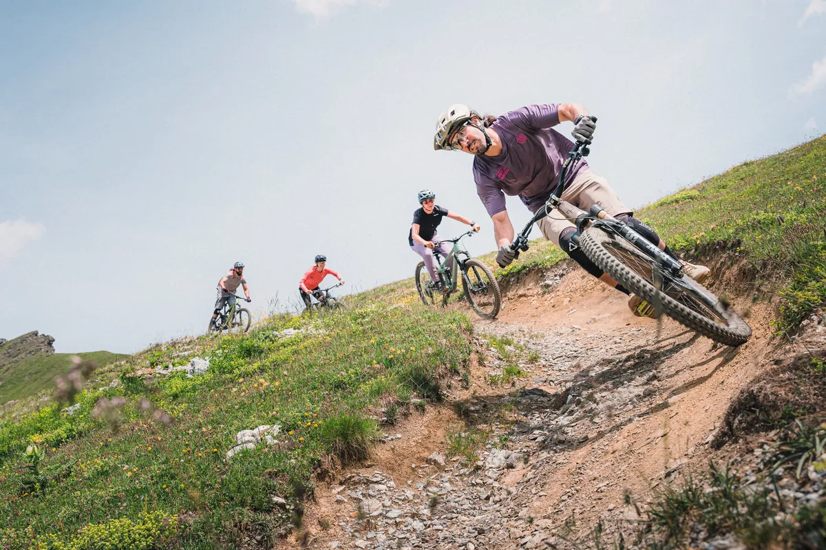 Mountainbiker legt sich mit seinem Trailbike in einen Anlieger auf einem Alpentrail, drei weitere Fahrende folgen am Hang.