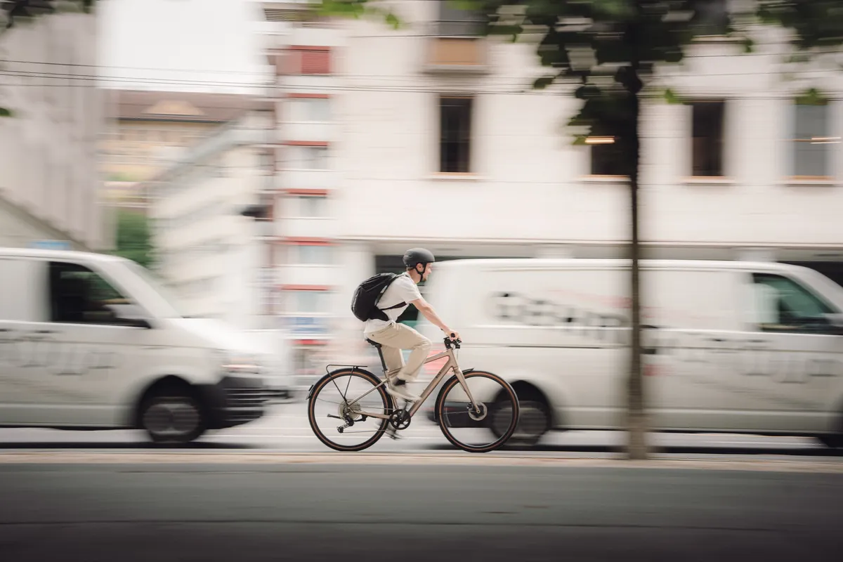 Pendler mit Helm und Rucksack fährt auf einem BIXS Urbanbike an parkierten Lieferwagen vorbei durch die Stadt