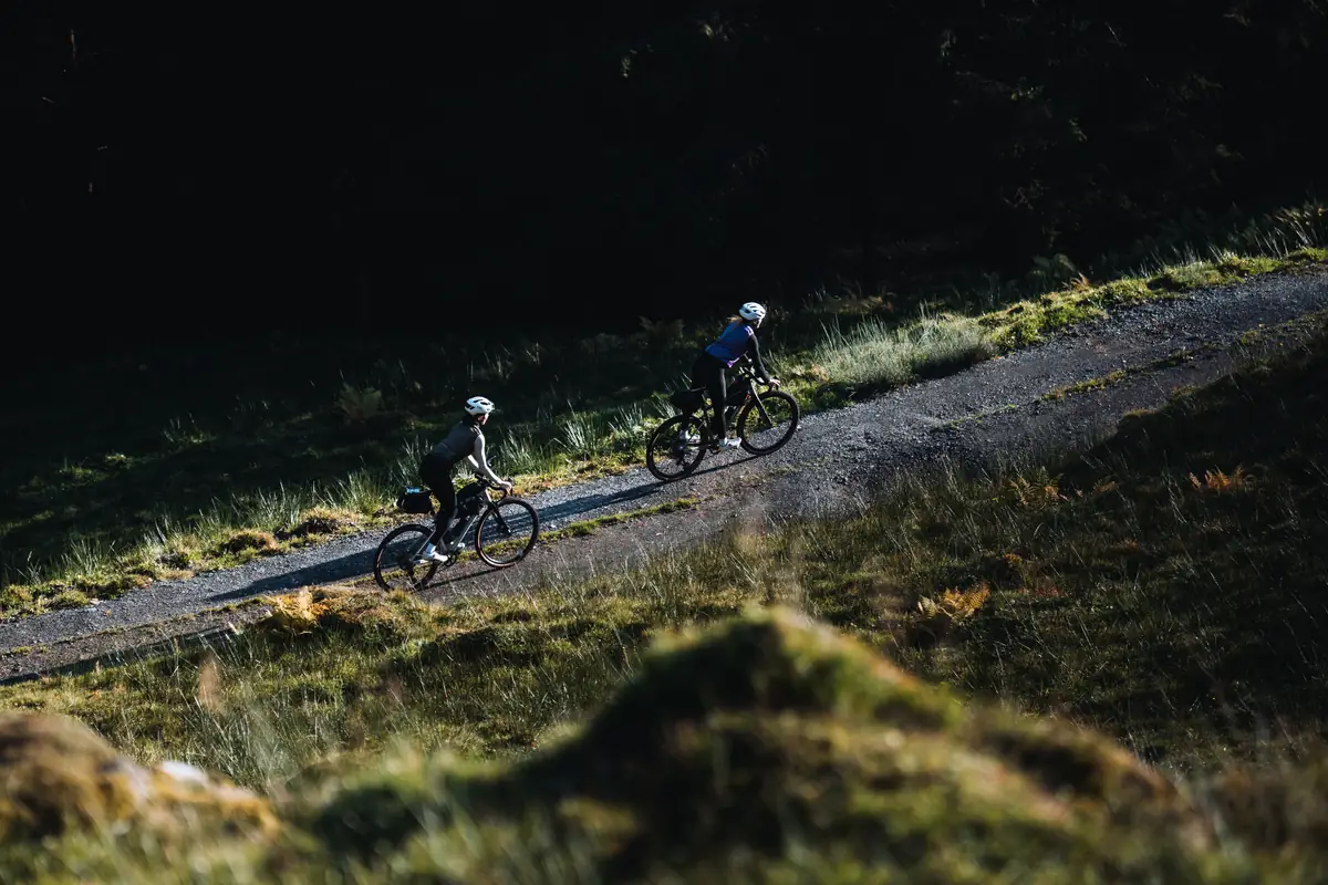 Zwei Fahrer auf beladenen Gravelbikes auf Schotterweg im Gegenlicht