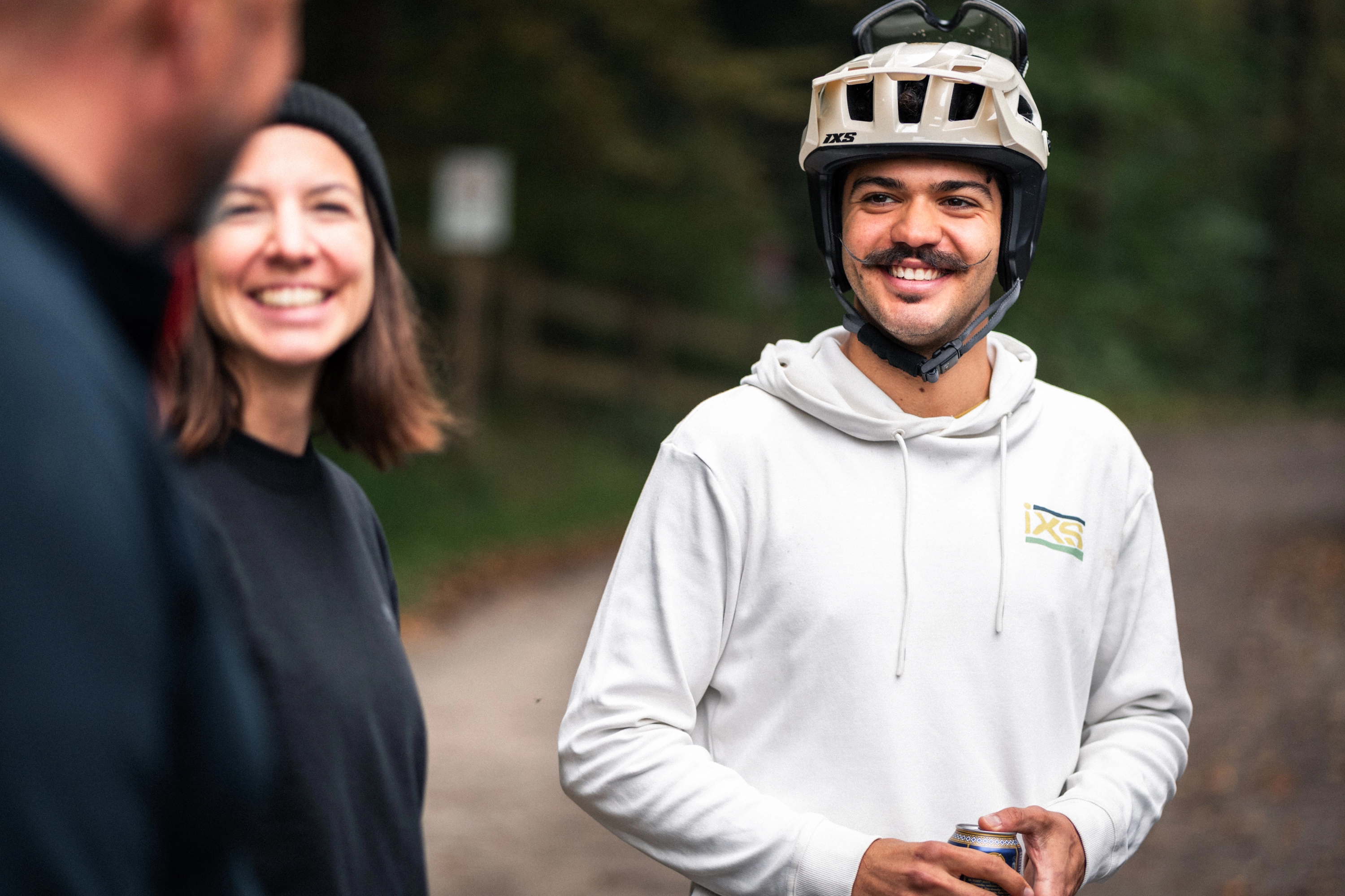 Lachender Mann mit Helm und IXS-Hoodie im Gespräch, Frau im Vordergrund unscharf