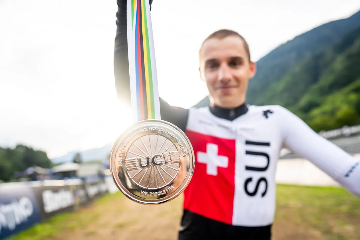 Luca Schätti hält seine WM-Medaille am Regenbogenband in die Kamera, im Hintergrund das Zielgelände