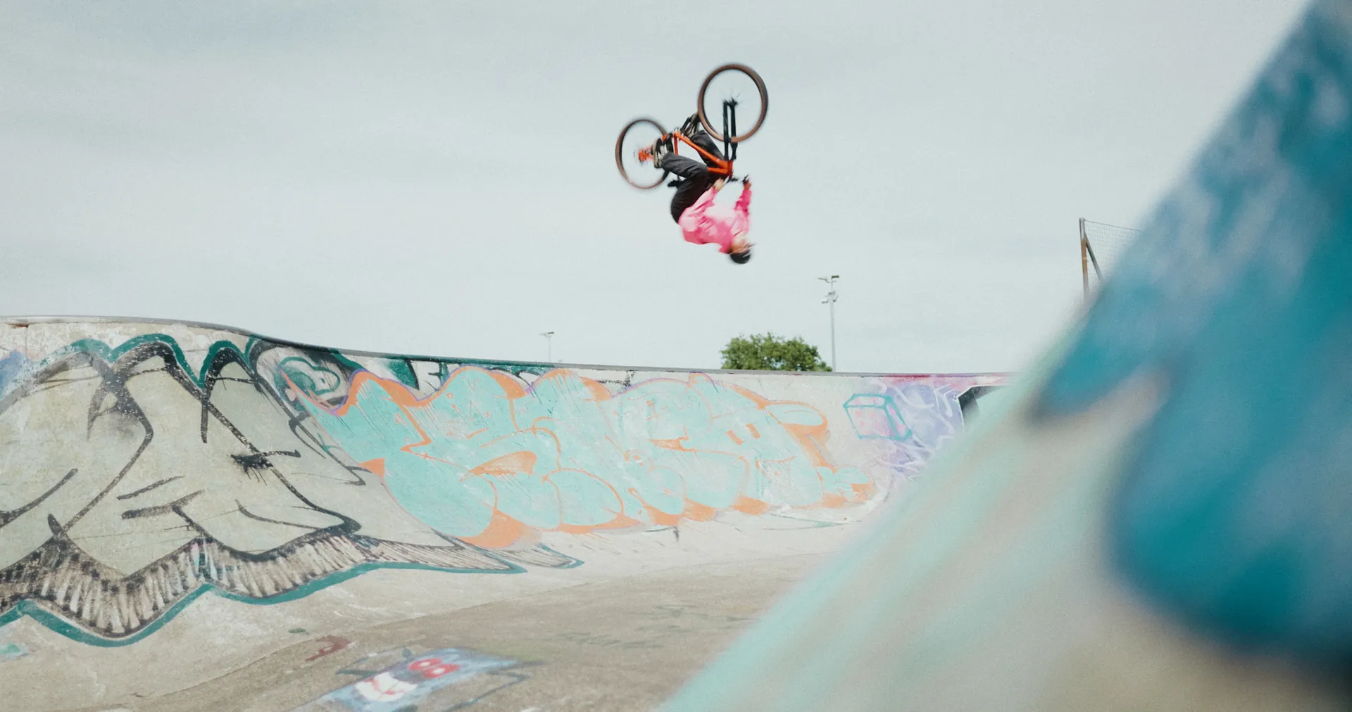 BIXS Fahrer im pinken Trikot vollführt einen Backflip mit dem Stomp über einer bemalten Skatepark-Bowl
