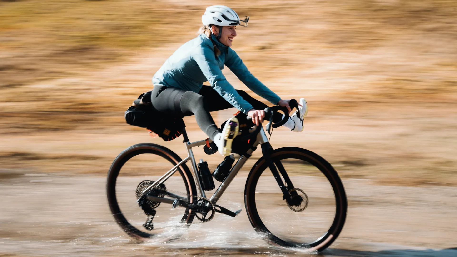 Bikerin fährt lachend mit bepacktem Gravelbike durch eine Wasserdurchfahrt, Bewegungsunschärfe im Hintergrund