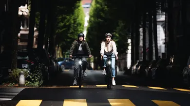 Zwei Fahrerinnen fahren mit BIXS District E-Bikes durch eine Baumallee in der Stadt