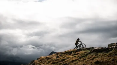 Fahrer schiebt BIXS Core-E Hardtail-E-Bike über Gipfelgrat über einem Wolkenmeer