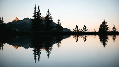 Zwei Mountainbiker als Silhouette am Bergsee im Abendlicht mit Spiegelung